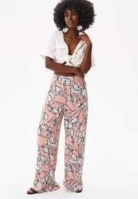 Różowe spodnie w szerokim kroju z kwiatowym wzorem, z czarno-białym motywem florystycznym, gładki materiał, wysoki stan, w zestawieniu z białą koszulą z węzłem i dodatkami.