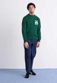 Sudadera verde con un logo redondo en el pecho, combinada con pantalones azul marino y zapatillas negras, de pie contra un fondo blanco.