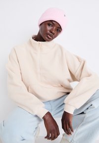 Fleece-Pullover in Creme mit Reißverschlusskragen und Fronttasche, kombiniert mit hellblauen Jeans und einer rosa Strickmütze. Weiche Textur und entspannte Passform.