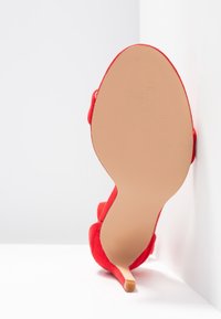 Chaussure à talon haut en daim rouge, posée talon vers le haut contre une surface blanche d'angle, montrant la semelle beige lisse et le petit embout du talon.