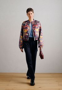 Veste bomber courte avec des motifs floraux et animaliers colorés ; associée à un pantalon à fines rayures sombres et un petit sac marron.