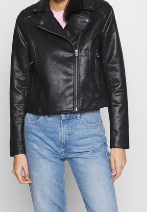 Nainen pukeutuneena mustaan keinonahkaiseen moottoripyörätakkiin, jossa on vetoketju edessä, yhdistettynä vaaleansinisiiin korkeavyötäröisiin farkkuihin.