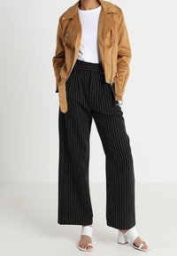 Veste en daim marron de motard, T-shirt blanc, pantalons larges à rayures noires, et sandales à talons blocs blanches. Textures lisses et design superposé.