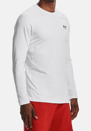 Homme portant un t-shirt blanc ajusté à manches longues avec un petit logo noir sur la poitrine et un short de sport rouge.