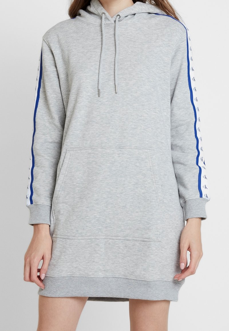 Femme portant une longue robe sweat à capuche grise avec une poche avant et des manches rayées bleues avec des logos "ck" répétés.