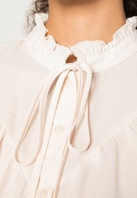 Blusa de color rosa claro con cuello con volantes, cierre de botones y lazada en el cuello. Tejido suave y liso con una textura sutil.