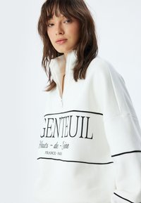 Sudadera blanca de media cremallera con diseño de texto negro que dice "GENTEUIL" y acentos. Tejido suave con un corte holgado y mangas anchas.