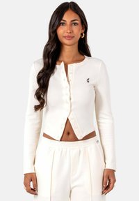 Witte cropped cardigan van zachte stof, met een knoopsluiting aan de voorkant en een klein embleem op de linkerborst. Gecombineerd met witte broeken.