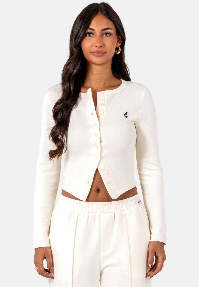 Witte cropped cardigan van zachte stof, met een knoopsluiting aan de voorkant en een klein embleem op de linkerborst. Gecombineerd met witte broeken.