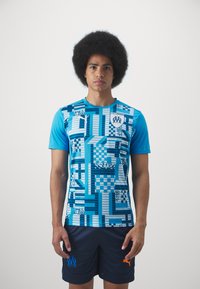 Cămașă sport cu mânecă scurtă de culoare albastră, cu modele geometrice, având nuanțe variate de albastru. Asortată cu pantaloni scurți negri, accente portocalii și logo-uri.