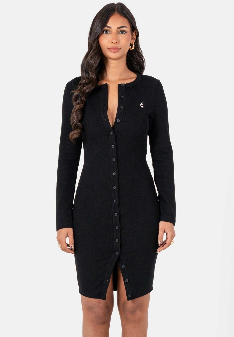 Vrouw met lang golvend haar, gekleed in een strakke zwarte blousejurk met lange mouwen, staande voor een eenvoudige witte achtergrond.