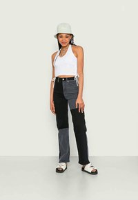 Jeans de retalhos em preto e cinza com uma cintura alta, combinados com uma blusa curta de alças brancas e sandálias brancas. Um chapéu de balde cinza claro completa o visual.