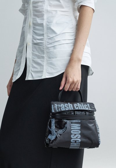 Henkilö, joka pitää mustaa Moschino-käsilaukkua, jossa on vaaleansiniset tekstit "trash chic!" yhdistettynä valkoiseen rypytettyyn paitaan ja mustiin hameisiin.