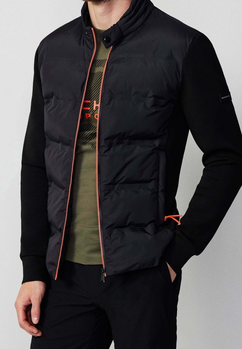 Homme portant une doudoune sans manches noire avec une fermeture éclair orange par-dessus un t-shirt vert à motif et une chemise noire à manches longues, main dans la poche.