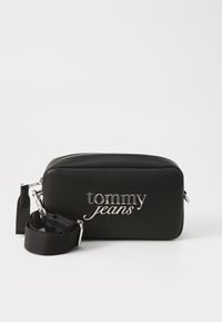Juodas sintetinės medžiagos šalinis krepšys su sidabriniu "tommy jeans" logotipu, užtrauktukas, reguliuojama petneša. Lygus tekstūros paviršius, stačiakampio forma, kompaktiškas dizainas.
