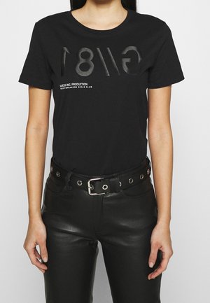 Femme portant un t-shirt noir avec une impression inversée discrète "GUESS 81" et un texte, associée à un pantalon en cuir noir et une ceinture noire avec des œillets argentés.
