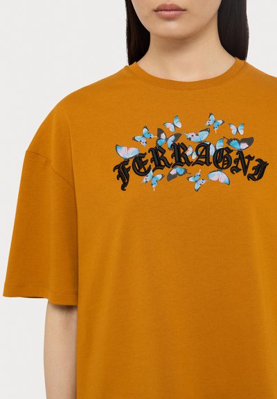 Πορτοκαλί βαμβακερό T-shirt με κοντά μανίκια, που διαθέτει κεντημένες πεταλούδες σε μπλε και μαύρο χρώμα και τη λέξη "FERRAGNI" σε στυλιζαρισμένη γραμματοσειρά.