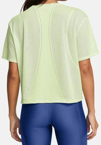 Mujer con una blusa de manga corta perforada de color verde claro y leggings azules ajustados, vista desde atrás.