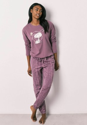Vrouw die een mauve Snoopy-sweatshirt en bijpassende gestructureerde loungebroek draagt, staat blootsvoets tegen een lichte, vlakke muur.