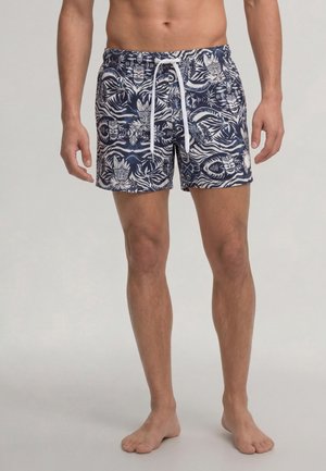 Mann trägt marineblaue und weiße Badeshorts mit tropischem Muster und einem weißen Kordelzug, steht barfuß auf neutralem Hintergrund.