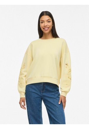 Noor naeratav noor naine, kandes kahvatukollast pusa lillemustriga varrukatel ja siniseid teksaseid, seistes valge tausta ees.