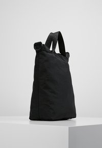 Borsa tote in morbido tessuto nero con robuste maniglie nere, poggiata su un piedistallo bianco contro uno sfondo grigio uniforme.