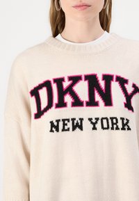 Suéter de malha em tom bege, com um decote canelado e um ousado logótipo em preto e rosa "DKNY NEW YORK" na frente. Textura suave.