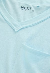 Világoskék V-nyakú póló mellzsebbel és "NEXT V-NECK TEE" felirat a gallér belsejében.