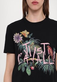Melnas kokvilnas T-krekls ar krāsainu ziedu un putnu grafiku, uz kuras ir teksts "JUST CAVALLI". Īsas piedurknes, apaļa kakla izgriezuma forma un detalizētas ilustrācijas.