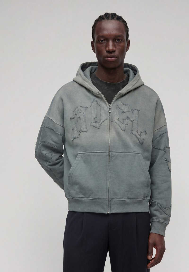 Homme aux cheveux tressés portant un sweat à capuche vert délavé zippé avec des lettres brodées en relief et un pantalon foncé, debout devant un fond uni.