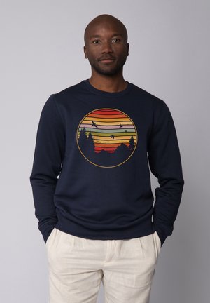 Marineblaue Sweatshirt mit einem kreisförmigen Grafikdesign, das übereinanderliegende Streifen in Rot, Orange und Gelb sowie Silhouetten von Bäumen und Vögeln zeigt.