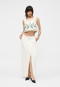 Γυναίκα με τατουάζ που φοράει ένα κρεμ crop top με κεντήματα λουλουδιών, φούστα με υψηλή μέση και άνοιγμα μπροστά σε λευκό χρώμα και μαύρα μυτερά παπούτσια.