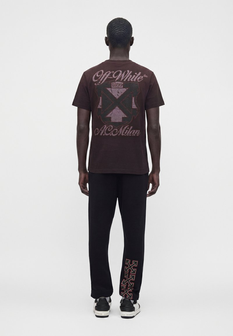 Homme portant un t-shirt Off-White foncé avec un texte graphique et un symbole dans le dos, pantalon noir avec des motifs de flèches rouges sur le bas de la jambe, et des chaussures noires et blanches.
