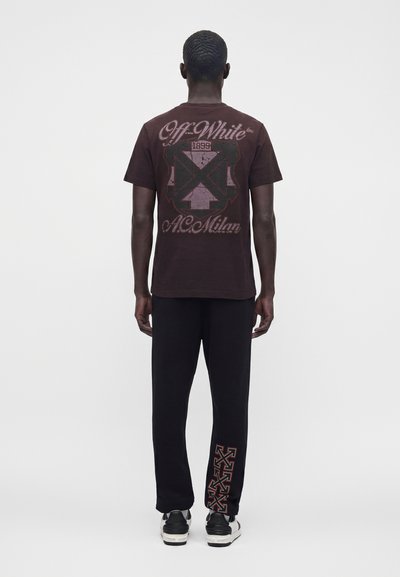 Άνδρας που φοράει σκούρο μπλουζάκι Off-White με γραφικό κείμενο και σύμβολο στην πλάτη, μαύρο παντελόνι με κόκκινες βέλη στο κάτω μέρος του ποδιού και μαύρα και λευκά παπούτσια.