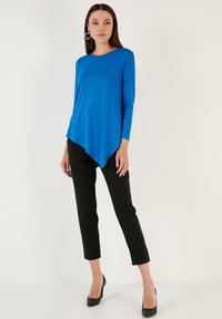Maglietta blu a maniche lunghe con orlo curvo, abbinata a pantaloni fitted neri e tacchi neri. Texture liscia, design minimalista e taglio asimmetrico.