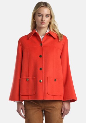 Moteris dėvi ryškiai oranžinę vilnonę striukę su sagomis ir kišenėmis su atvartais, derintą su rudomis kelnėmis, stovinti priešais paprastą foną.