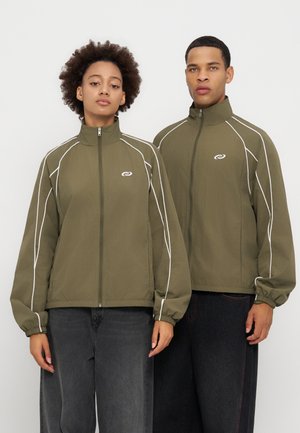 Deux personnes portent des vestes zippées vert olive assorties avec un liseré blanc et un petit logo sur la poitrine, debout devant un fond uni.