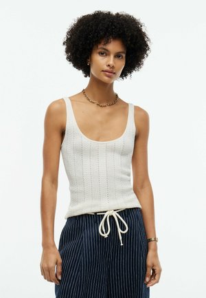 Vrouw met krullend haar die een witte gebreide tanktop draagt, marineblauwe broek met fijne strepen en trekkoord, een gouden ketting en een polshorloge.