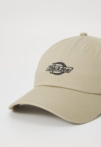 Beež baseballi müts, mis on valmistatud tekstureeritud kangast, reguleeritava rihmaga, esiosas musta tikandiga logo.