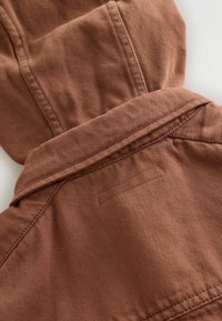 Veste à capuche marron en tissu texturé. Comprend un col, des accents cousus et une pièce rectangulaire au dos pour le marquage.