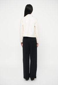 Γυναίκα με μακριά μαύρα μαλλιά, ντυμένη με άσπρο σακάκι και μαύρο παντελόνι φαρδύ, στέκεται μπροστά σε ένα απλό λευκό φόντο.