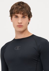 Fekete, hosszú ujjú rash guard rugalmas anyagból, mellkason elhelyezett logóval és lapos varrással a kényelem és tartósság érdekében.