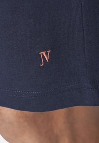 Marineblaue, gerippte Stoff mit kleinen, orangefarbenen, gestickten "JV"-Initialen nahe dem Saum, getragen über gebräunter Haut.