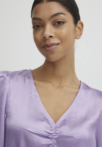 Blouse en satin lavande avec un col en V et des détails froncés au centre. Le tissu a une texture lisse et brillante avec des manches bouffantes.