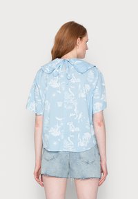 Blusa de manga curta azul clara com estampado botânico branco, apresentando um grande colarinho e detalhe de laço nas costas. Tecido macio e texturizado.