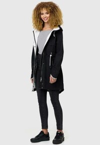 Black waterproof jacket with a white-lined hood, front zipper, drawstring waist, and multiple pockets, paired with black jeans and sneakers.