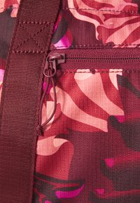 Gros plan sur un tissu texturé avec des motifs floraux roses et bordeaux, avec une poche zippée et une bandoulière robuste visibles.