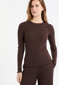 Braunes, geripptes Langarmshirt mit rundem Ausschnitt und dekorativen Knöpfen auf der Schulter; verfügt über eine lockere Passform und Rüschen am Saum.