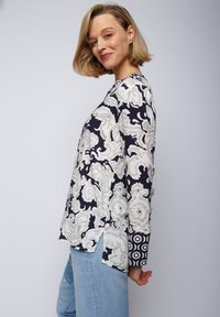 Musterbluse mit langen Ärmeln in Marineblau und Creme, mit floralen Motiven und kreisförmigen Akzenten an den Ärmeln. Weicher Stoff und lockere Passform.