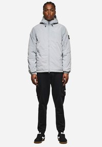 Giacca puffer grigia chiara con cappuccio, chiusura con zip e polsini elasticizzati, abbinata a pantaloni cargo neri con tasche laterali e sneakers nere.